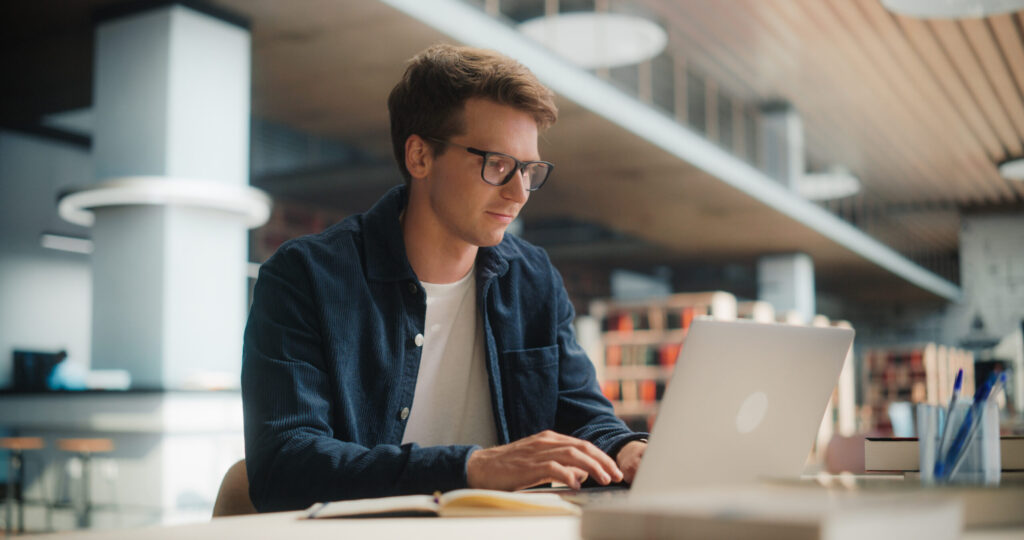 Concentrated Young Caucasian Male Student Working on His Laptop in a Modern Library. Man Engaged in Academic Research With Books Around, Focused on Screen